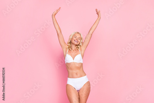 Photo portrait of attractive elderly woman raise hands enjoy freedom wear trendy white lingerie isolated on pink color background