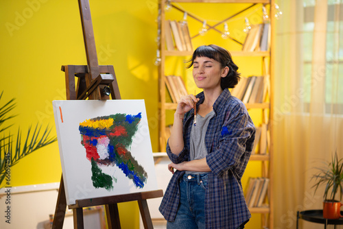 A woman is rustling through many different types of brushes while painting something astonishing on a large canvas that is held up by an easel