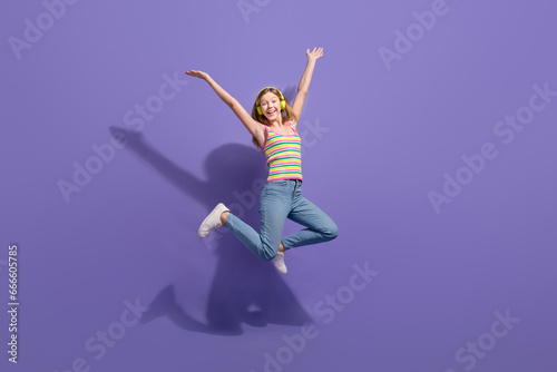 Full size photo of pretty teenager blonde girl jump raise hands excited wear trendy striped outfit isolated on purple color background
