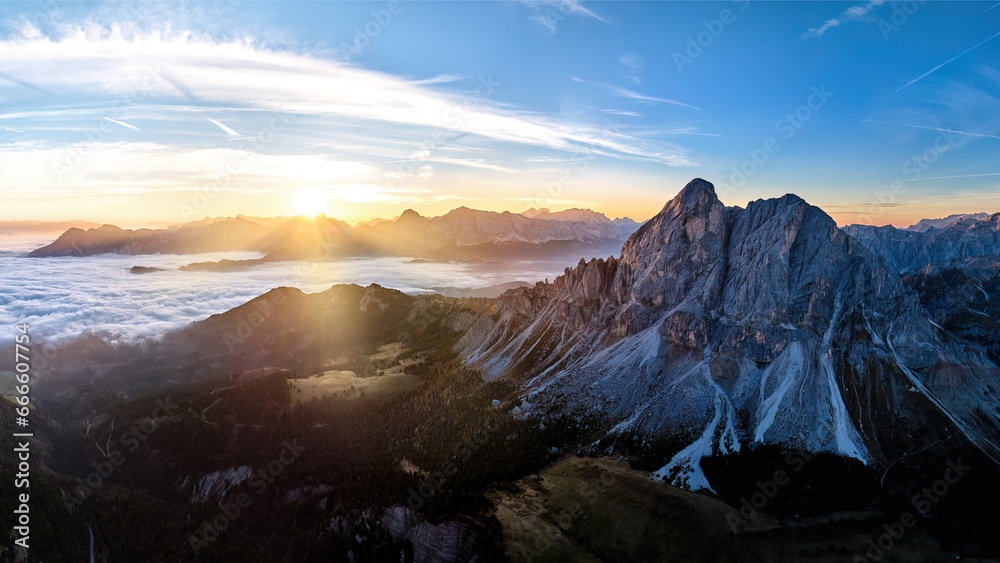 Sunrise over the Sass de Puta mountain peak at Passo delle Erbe pass against the Dolomite peaks in the background, inverse cloud cover in the valley, sun rays. Aerial drone mountainscape panorama.
