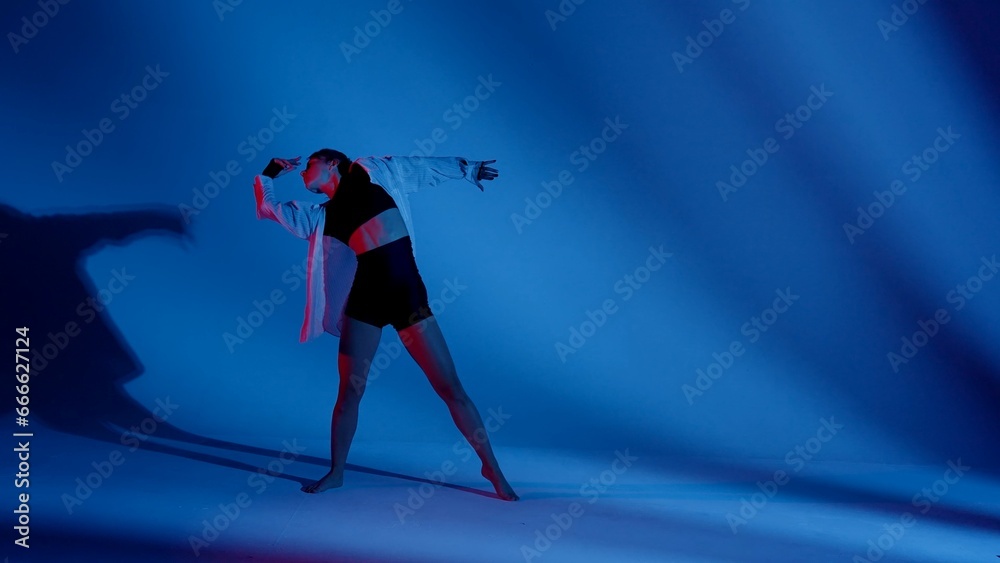 Young woman wearing a top, shorts and a shirt performing contemporary dance in studio. Neon blue and red color scheme, shadowed background. Full length.