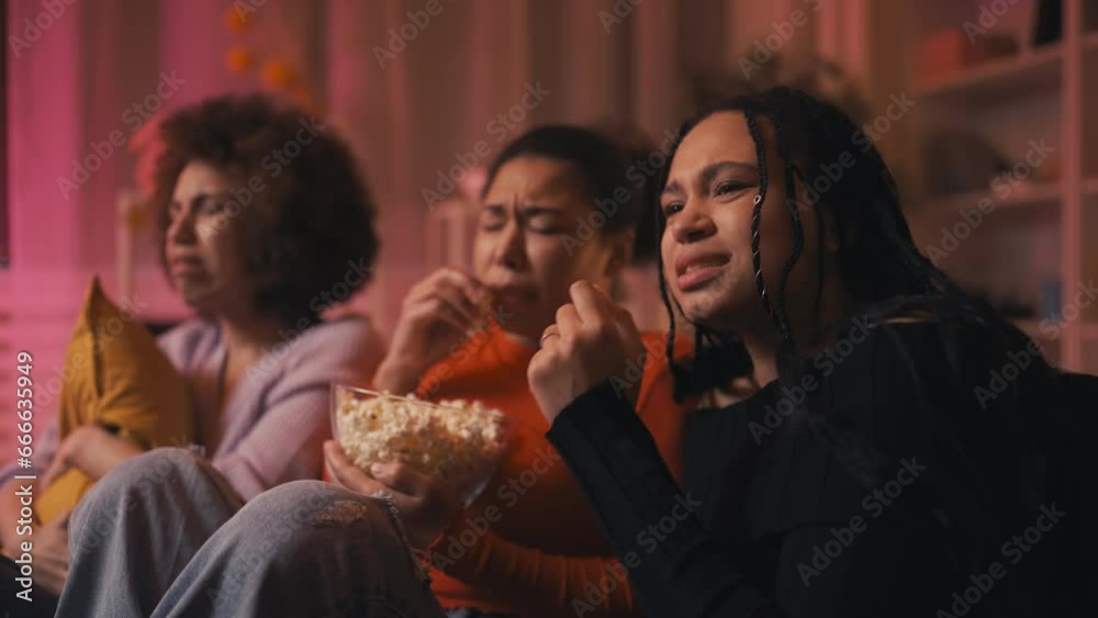 Emotional African American young women watching horror movie, eating popcorn