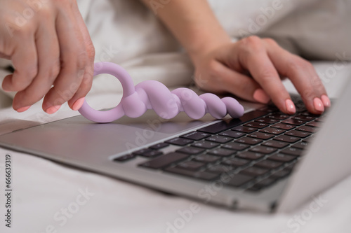 Woman holding lilac anal beads next to laptop while lying on bed. 