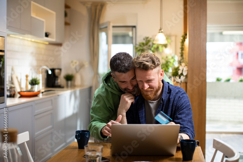 Happy young gay couple shopping online with a credit card on the laptop at home