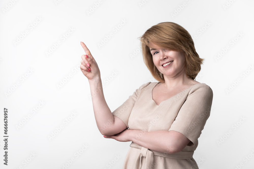 Blonde woman mature adult wearing casual beige dress looking at camera with pleasant smile, standing on white background. Female finger is raised to image advertisement. Part of series. Copy space Blonde woman mature adult wearing casual beige dress looking at camera with pleasant smile, standing on white background. Female finger is raised to image advertisement. Part of series. Copy space