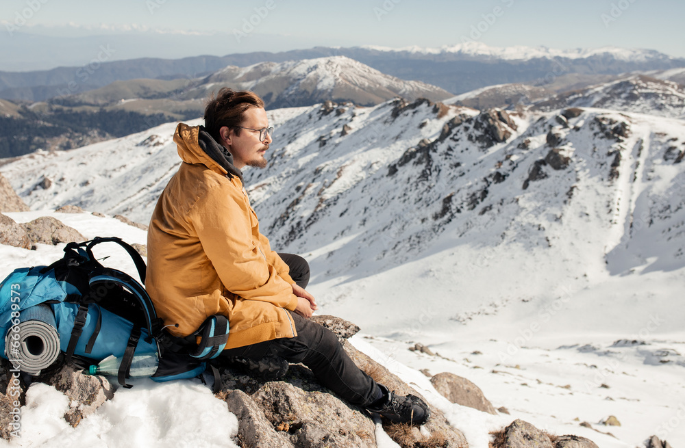 Portrait of a man in the mountains: a man sits on a peak with a backpack behind him against a mountain backdrop. High-quality photo for website design, postcards, banners, and travel product advertisi