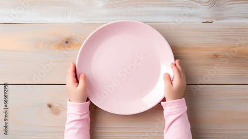 A baby is seen waiting for breakfast with an empty pink plate and a child's hand against a flat lay background made of white wooden boards at the top of the image.