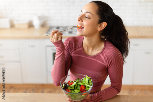 Sporty lady eats healthy veggie salad in modern home kitchen