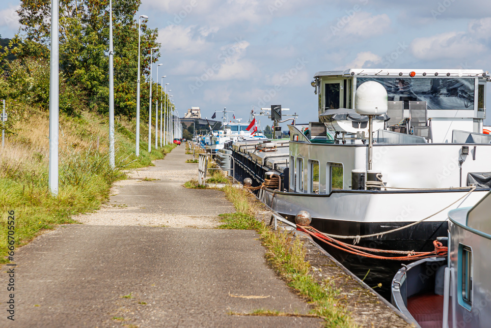 Naklejka premium Pedestrian path along canal with cargo ship anchored at pier on coast, lane lock, street lamps post in line, green trees, sunny day with hazy blue sky in Ternaaien, Belgium