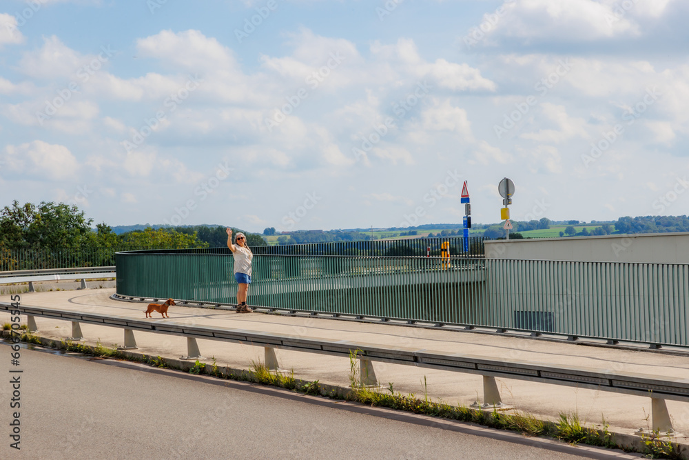 Rural road and pedestrian path and bicycles at Lanaye locks, smiling senior female tourist standing with her dog next to a metal fence waving with raised hand, sunny summer day in Ternaaien, Belgium