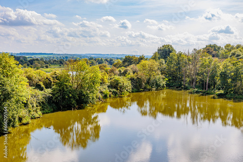 Part of the Maas river surrounded by green leafy trees, Dutch countryside and horizont in misty background, seen from Lanaye lock, sunny day, blue sky and white clouds, Ternaaien, Vise, Belgium