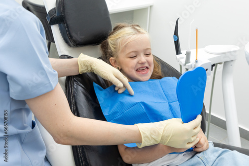 Lovely little girl looking in the mirror at her teeth after teeth surgery in a pediatric stomatology. Happy kid after teeth examination.