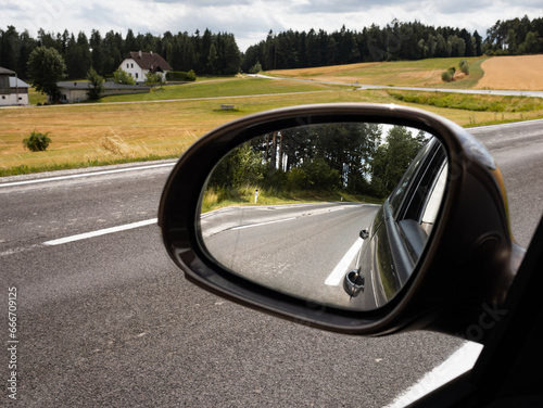 View into the car side mirror on the left side. The street in the rural area is empty. The blind spot is not visible in the rearview mirror.