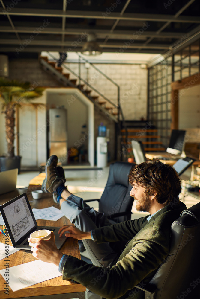 Young Caucasian male designer drinking coffee while working on the laptop in a startup company office Young Caucasian male designer drinking coffee while working on the laptop in a startup company office