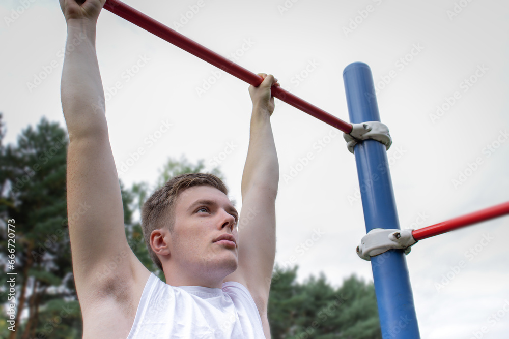 A cute young man is doing exercises on a horizontal bar at the stadium