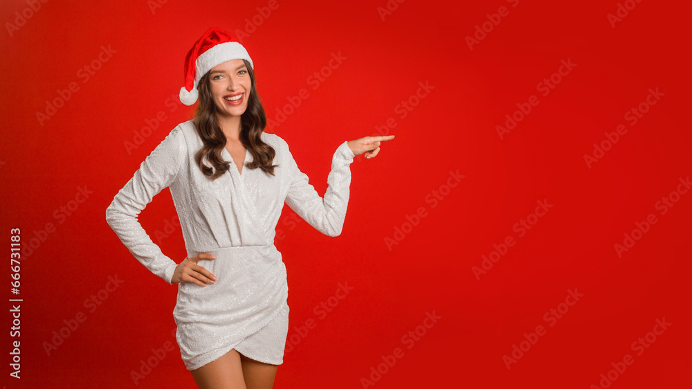 Woman in Santa hat and dress pointing finger, red background