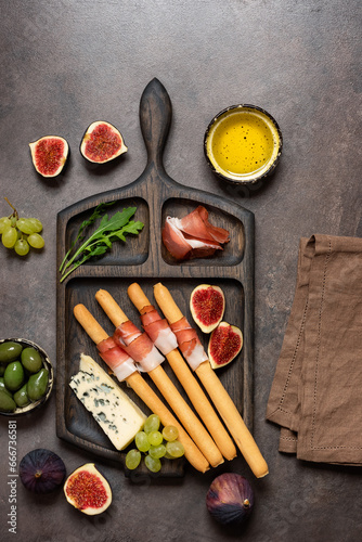Bread sticks grissini with prosciutto ham , blue cheese, figs, grape and olives on dark rustic background, closeup. Italian antipasti . Top view, flat lay, vertical