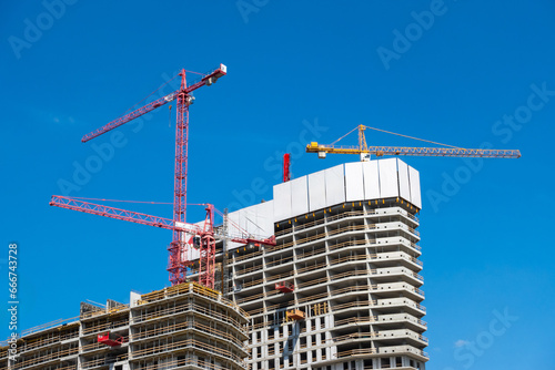 Obraz na płótnie Construction cranes on the construction site of a concrete apartment building. The building and cranes against the blue sky. Photo taken on a sunny day. w salonie