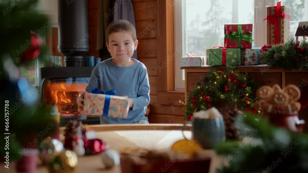 little joyful boy chooses wrapped box, sits on floor and put present on table look at it and shakes it smiling. christmas decorations, fir tree branches, flashing lights and fireplace on background little joyful boy chooses wrapped box, sits on floor and put present on table look at it and shakes it smiling. christmas decorations, fir tree branches, flashing lights and fireplace on background