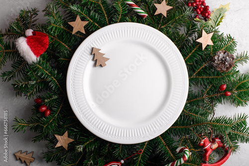 Christmas table setting with white ceramic plate on natural fir branches background. Christmas festive decoration serving for Christmas dinner, flat lay, top view, greeting card template