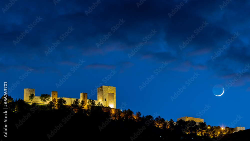 Santa Catalina Castle in Jaén (Spain) illuminated at night, with a cloudy blue sky, and the waxing crescent moon on the right side of the image
