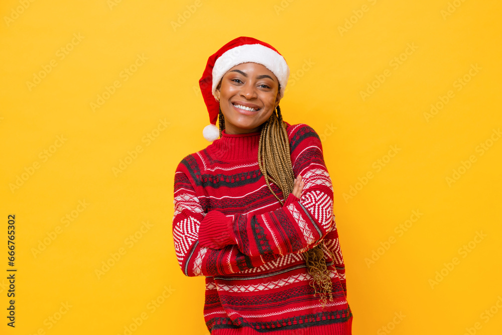African Amercian woman in red Christmas sweater and santa hat smiling with arms crossed in yellow color isolated background studio shot African Amercian woman in red Christmas sweater and santa hat smiling with arms crossed in yellow color isolated background studio shot