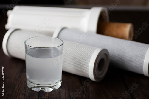 Used cartridges for a water purification filter and a glass of water on a dark wooden table. Soft focus.