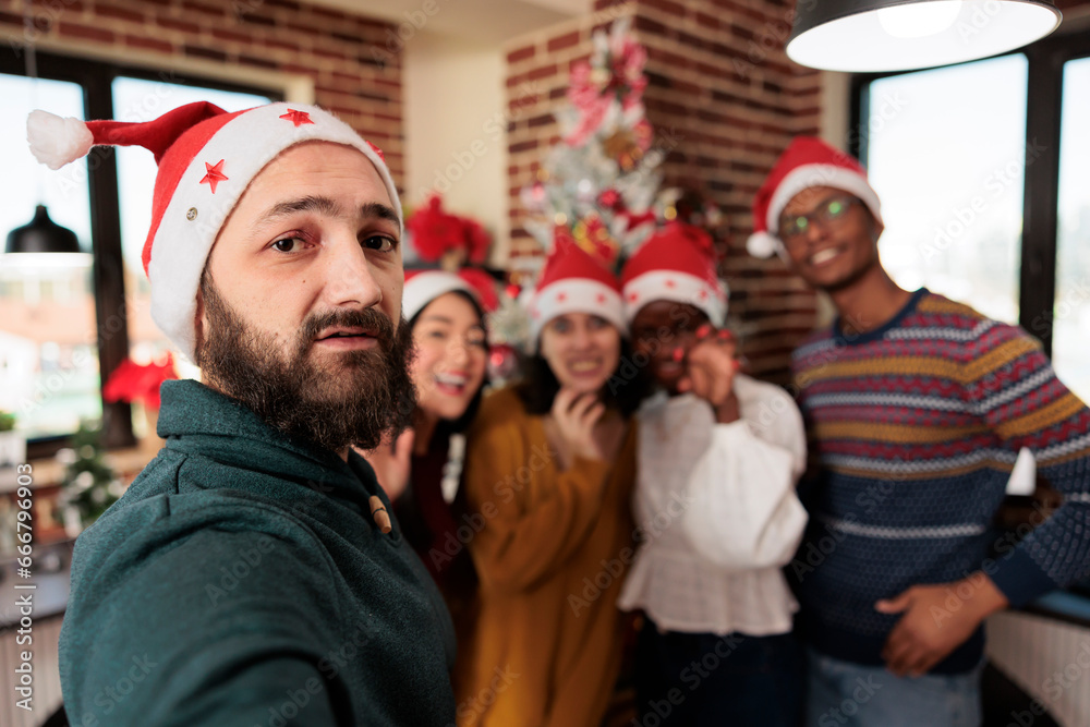 Fototapeta premium Diverse workers taking smartphone selfie at christmas corporate party in decorated office. Colleagues in santa hats posing for mobile phone photo while celebrating new year at workplace