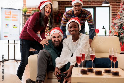 Excited employees holding gamepads and enjoying video game in festive decorated office on xmas eve. Cheerful diverse company coworkers fighting in videogame at new year holiday party