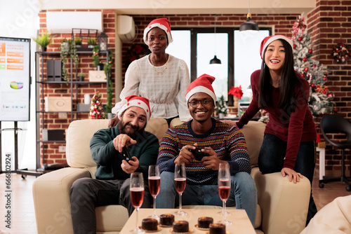 Startup diverse employees team enjoying festive christmas celebration in office. Colleagues drinking and playing video games with joysticks at winter seasonal corporate party