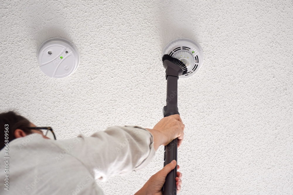 Woman vacuuming smoke and carbon monoxide detectors on a ceiling of a domestic room - obrazy, fototapety, plakaty 