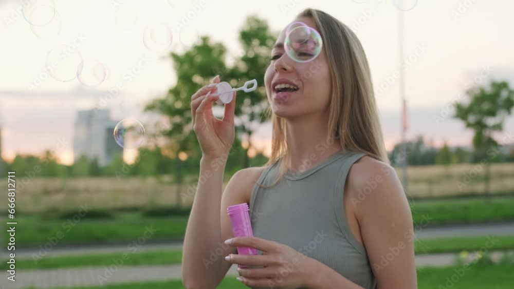 Happy young woman with long loose hair blows soap bubbles laughing in city garden. Smiling lady blows bubbles having fun in park in evening Happy young woman with long loose hair blows soap bubbles laughing in city garden. Smiling lady blows bubbles having fun in park in evening