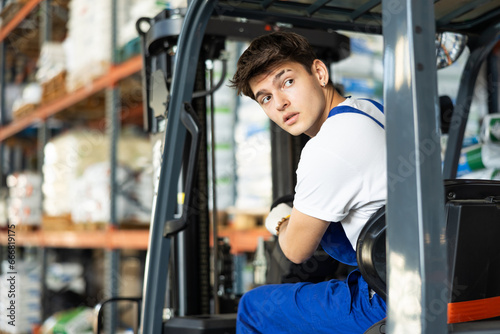Guy works on fork lift loader in construction rack area warehouse store repair materials. Young male employee looks back, chooses safe route for movement of equipment mechanism