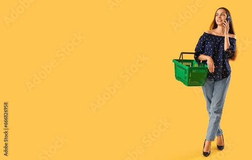 Young woman holding empty shopping basket and talking by phone on yellow background with space for text