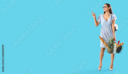 Young woman holding shopping basket full of products and pointing at something on light blue background with space for text