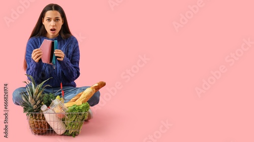 Shocked young woman with empty wallet sitting near shopping basket full of products on pink background with space for text