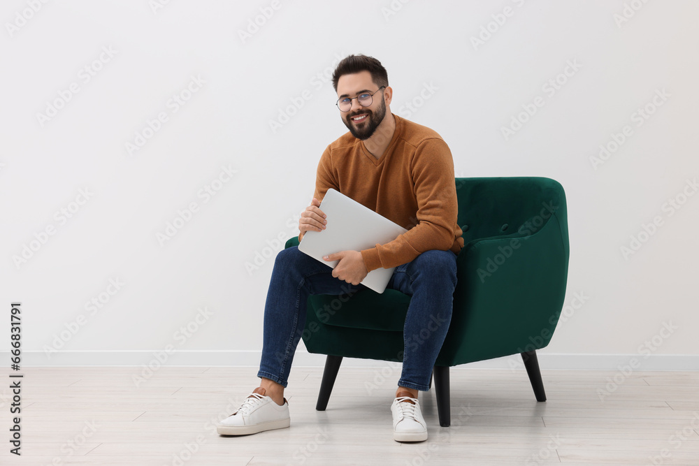 Handsome man with laptop sitting in armchair near white wall indoors, space for text