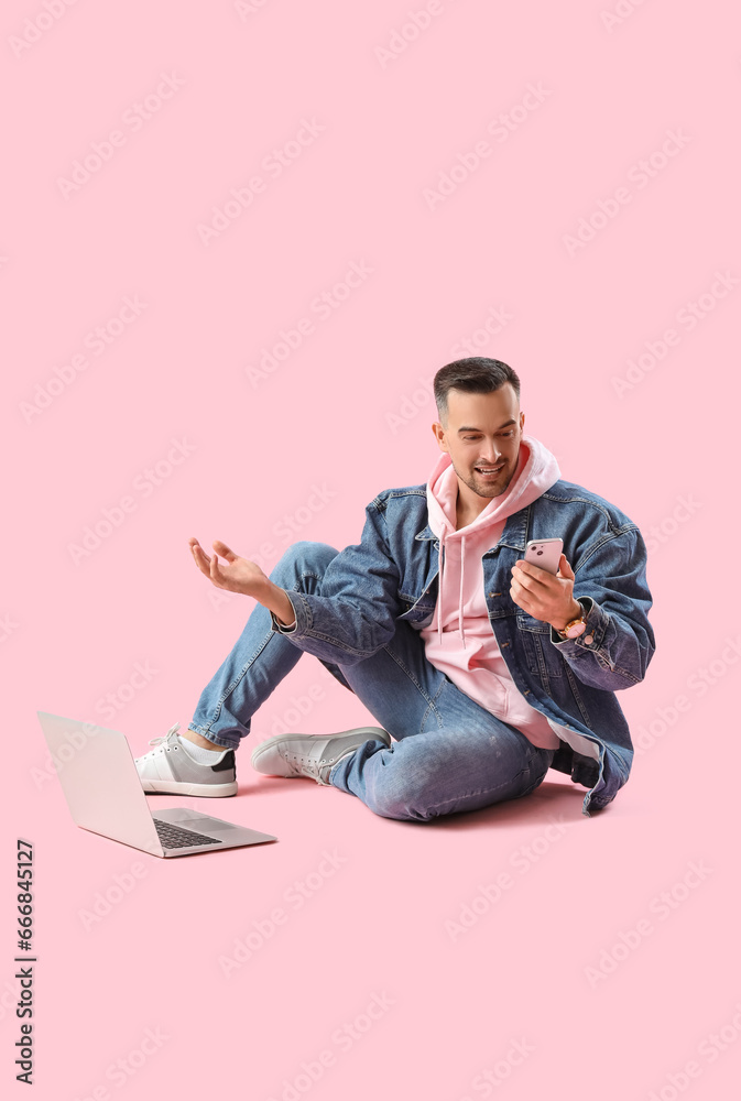 Happy young man with mobile phone and laptop sitting on pink background Happy young man with mobile phone and laptop sitting on pink background