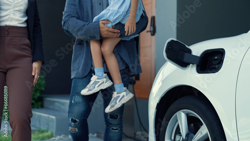 Progressive young parents and daughter living in a home with an electric vehicle and electronic vehicle charging station. Green and clean energy from electric vehicles for healthy environment. photo