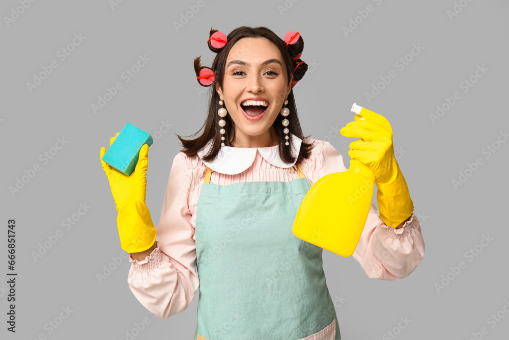 Portrait of happy young housewife in apron with sponge and detergent on grey background Portrait of happy young housewife in apron with sponge and detergent on grey background