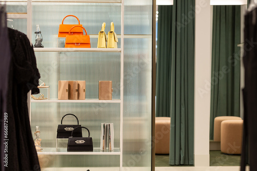An assortment of shoes and handbags are seen displayed on a white shelving unit in a trendy retail store. Fluted glass can be seen behind the items.