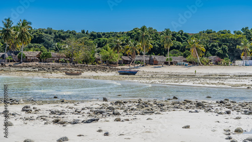 Low tide in the ocean. Stones are scattered on the exposed seabed. Wooden boats on the sand. On the shore, in the thickets of palm trees, Malagasy huts, drying laundry, tiny silhouettes of people © Вера 