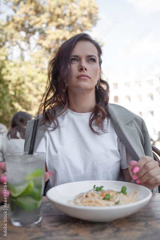 A beautiful woman sit in cafe with cup of drink and pasta eagainst  street of the city. A business woman looking sidesways. the female wearing the style basic outfit,  black trousers