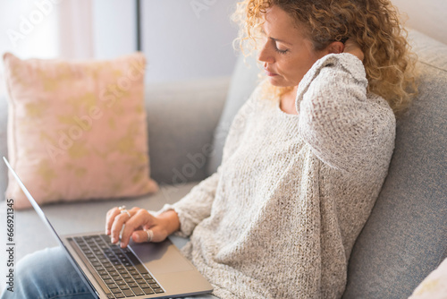 One woman sitting at home on the sofa using laptop tired and relaxed. Wireless computer work people. Adult female surfing the web alone indoor. Touching neck for pain and bad posture unhealthy