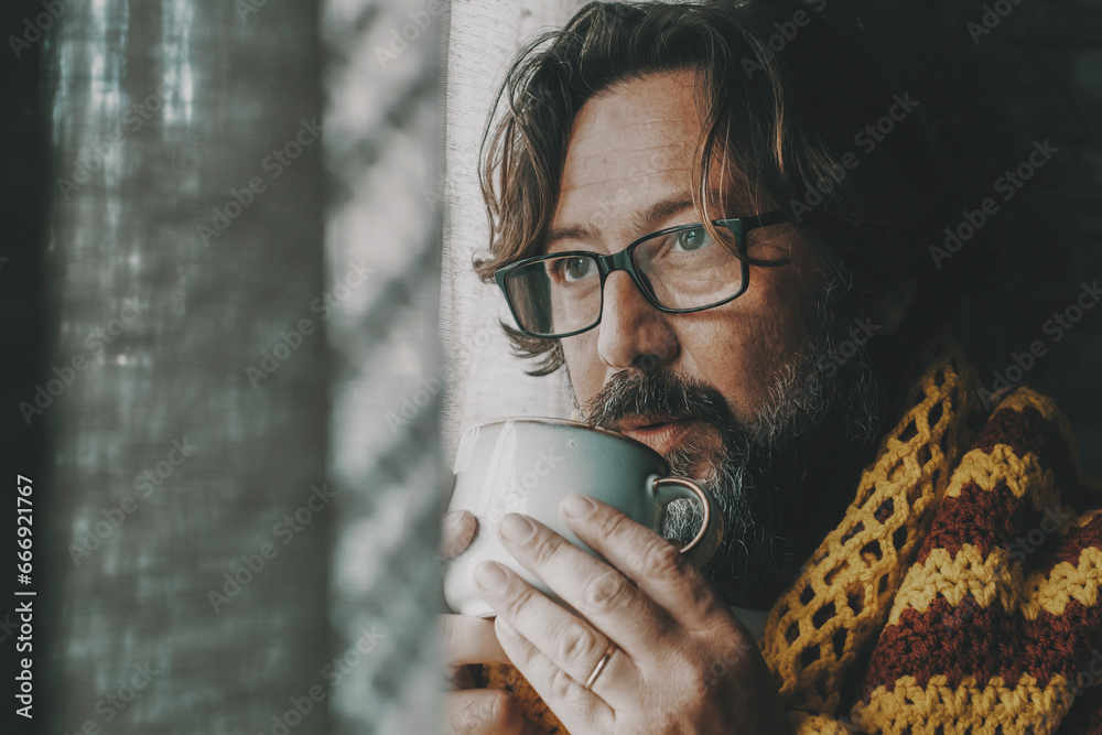 Side portrait of unhealthy man unhappy at home drinking medicine or coffee near the window looking outside. Cold interior temperature concept. Gas energy crisis problems. Flu and cold health people - obrazy, fototapety, plakaty 