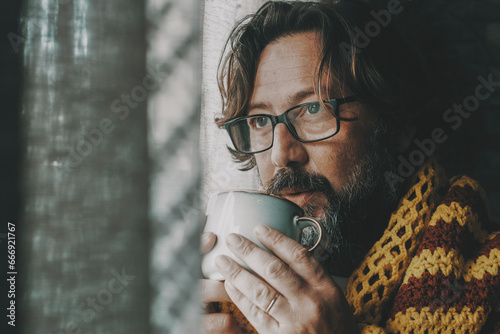 Side portrait of unhealthy man unhappy at home drinking medicine or coffee near the window looking outside. Cold interior temperature concept. Gas energy crisis problems. Flu and cold health people photo