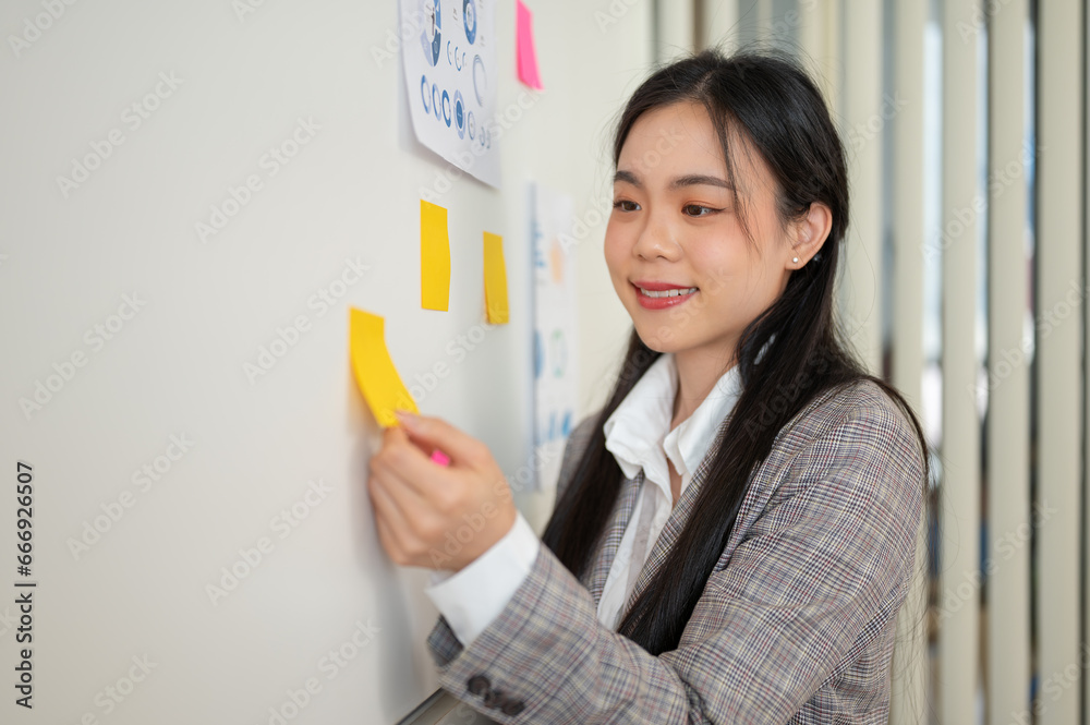 An Asian businesswoman is attaching a sticky note to the wall and sharing her idea with her team. An Asian businesswoman is attaching a sticky note to the wall and sharing her idea with her team.