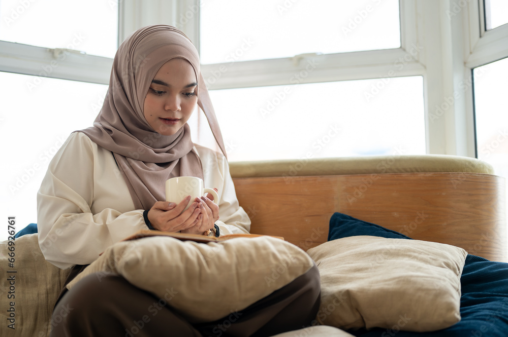 A focused Asian-Muslim woman is enjoying her coffee while reading a book in the living room.