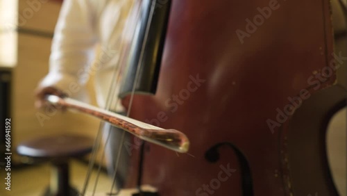 extreme close up of weary used scratched cello bass being played slowly by a professional musician inside building with wooden walls little round seat next to him man wearing white shirt grey pants photo