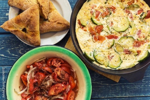 Fried omelette with zucchini, leeks and tomatoes served in a frying pan with marinated tomatoes with onions and pies on a rustic wooden background.